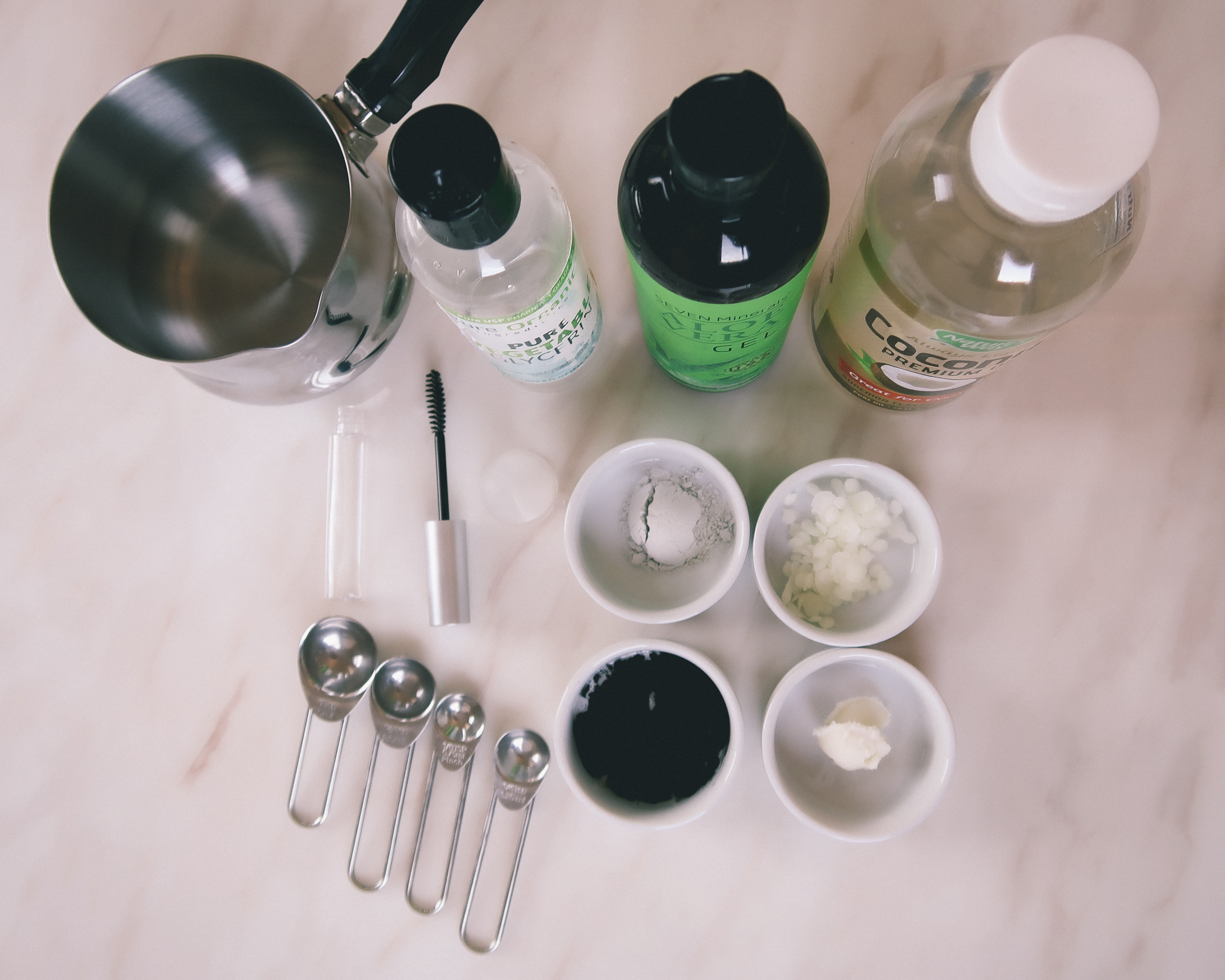 clean mascara ingredients laid out in small ramekins and bottles on white marble along with teaspoons, empty mascara tube and small melting pot
