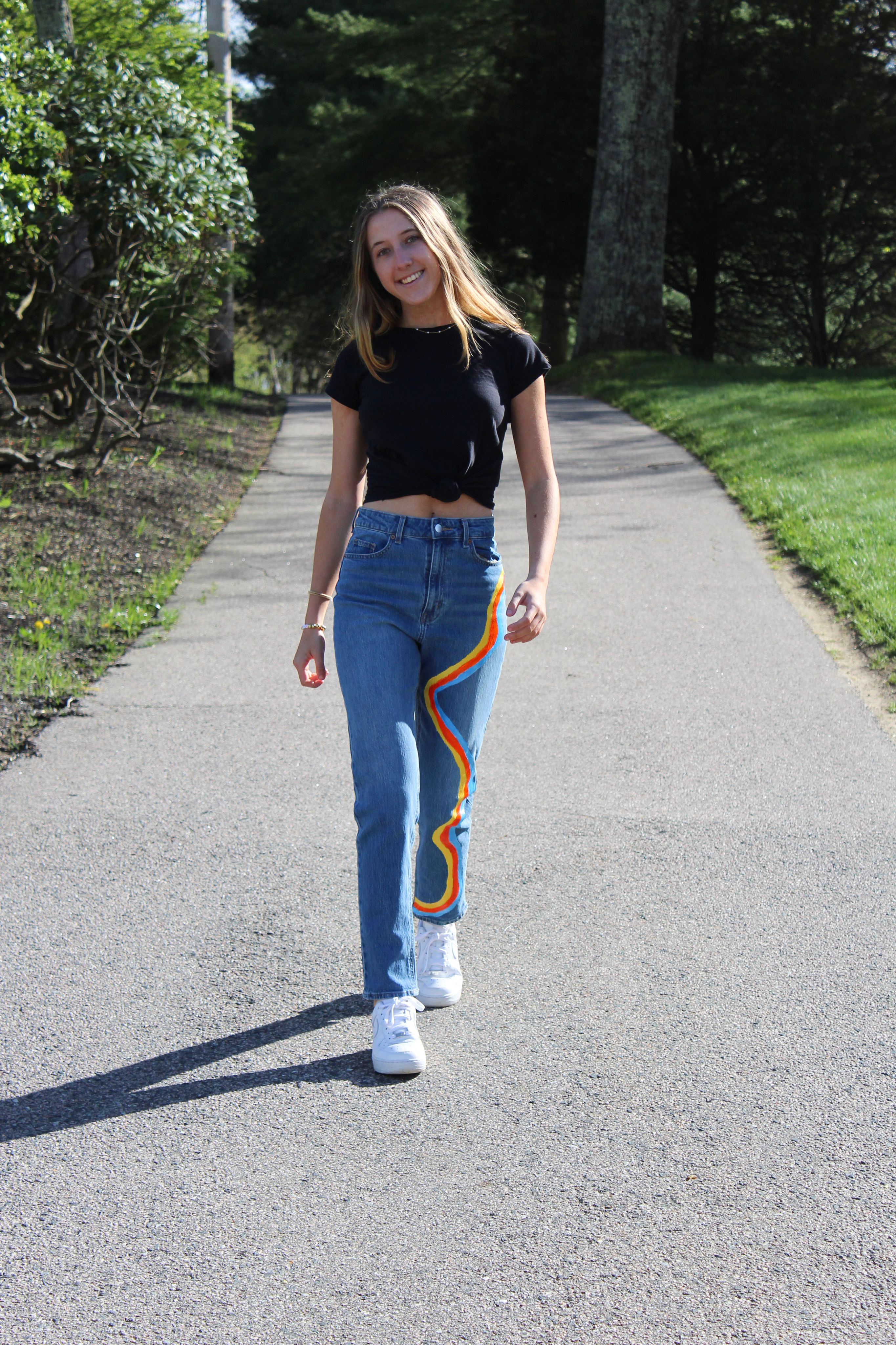 Girl walking down path in black t-shirt and jeans painted with stripes of color down one leg.